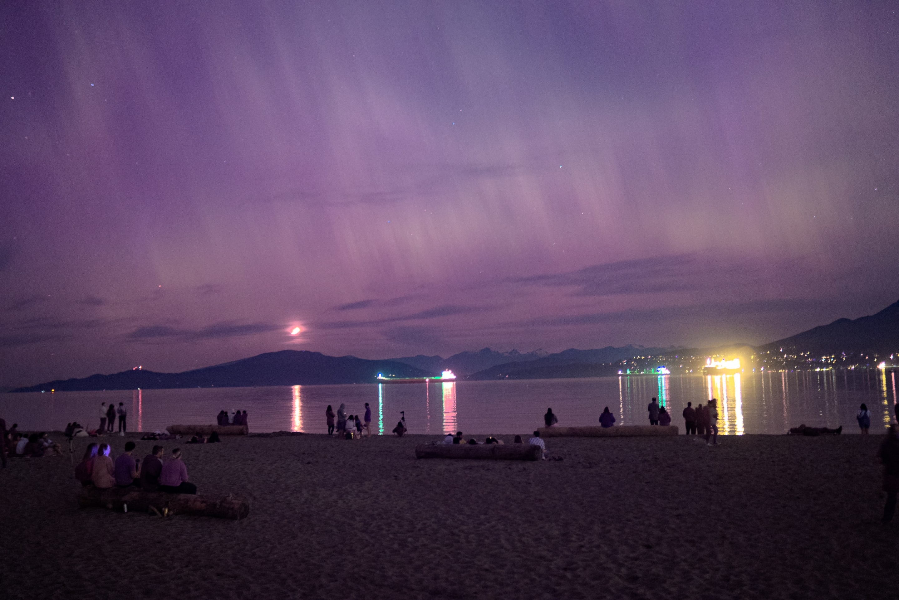 A nighttime beach scene under a vibrant purple sky. Silhouettes of people on the beach contrast with the lights reflecting on the water.