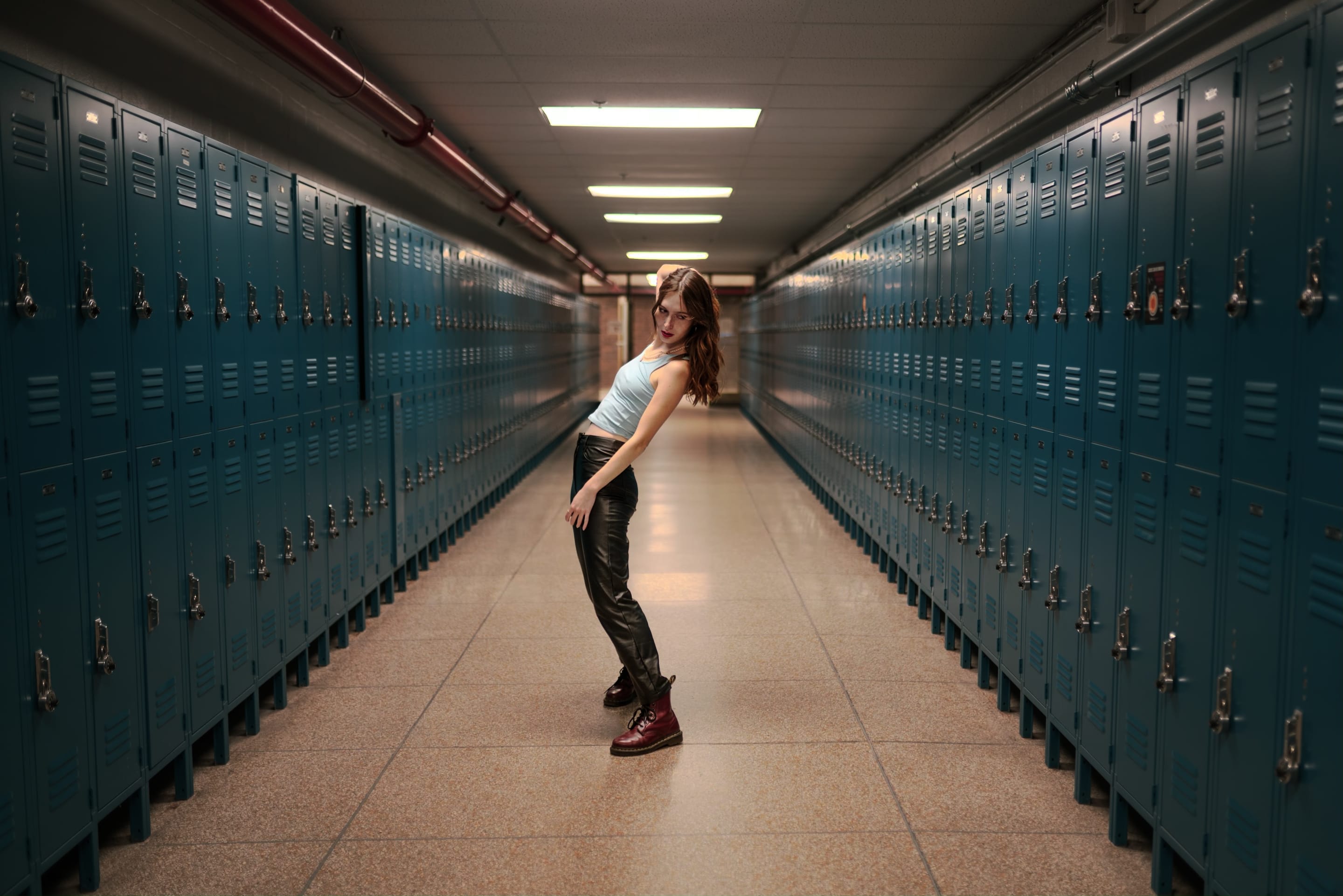 A woman poses in a school hallway, leaning back dramatically. The scene is framed by rows of teal lockers, leading the eye into the distance.