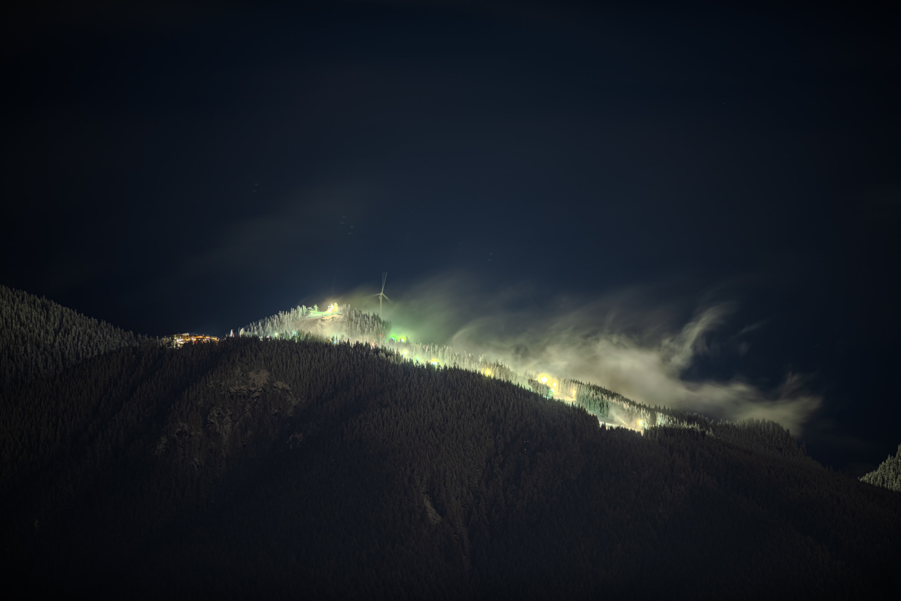 A nighttime landscape shows a mountain range with illuminated slopes and a cloudy sky. The scene is illuminated by lights scattered across the mountain.