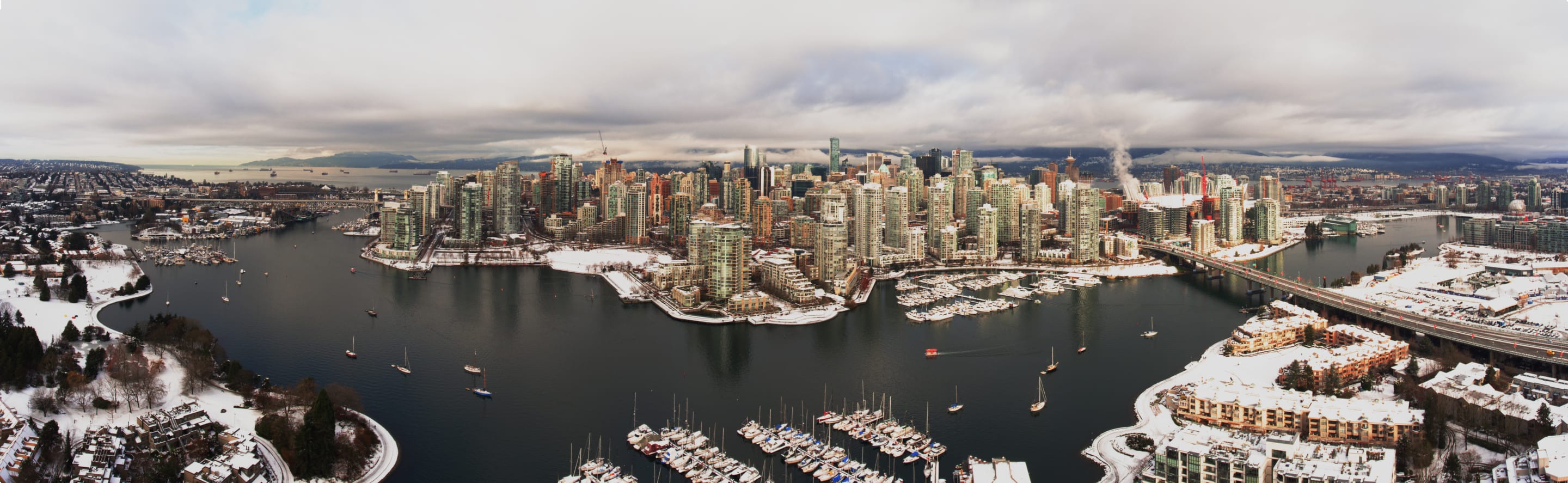 An aerial panoramic view of a city skyline on a cloudy day. The city has tall buildings and is situated near a body of water with boats.