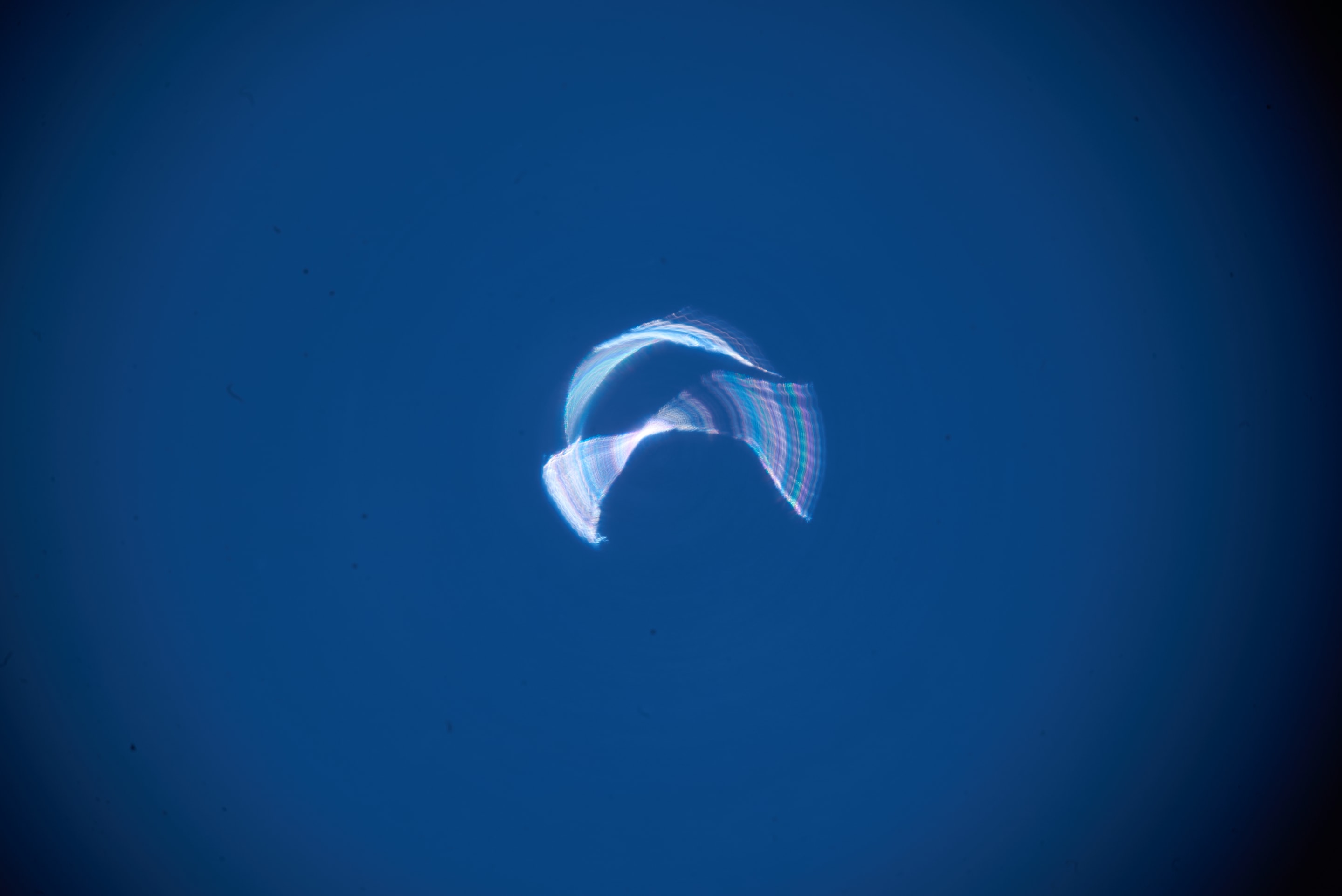 A swirling, luminous ribbon of light, composed of iridescent blues, purples, and whites, drifts across a deep blue expanse. The abstract form suggests movement and ethereal energy.