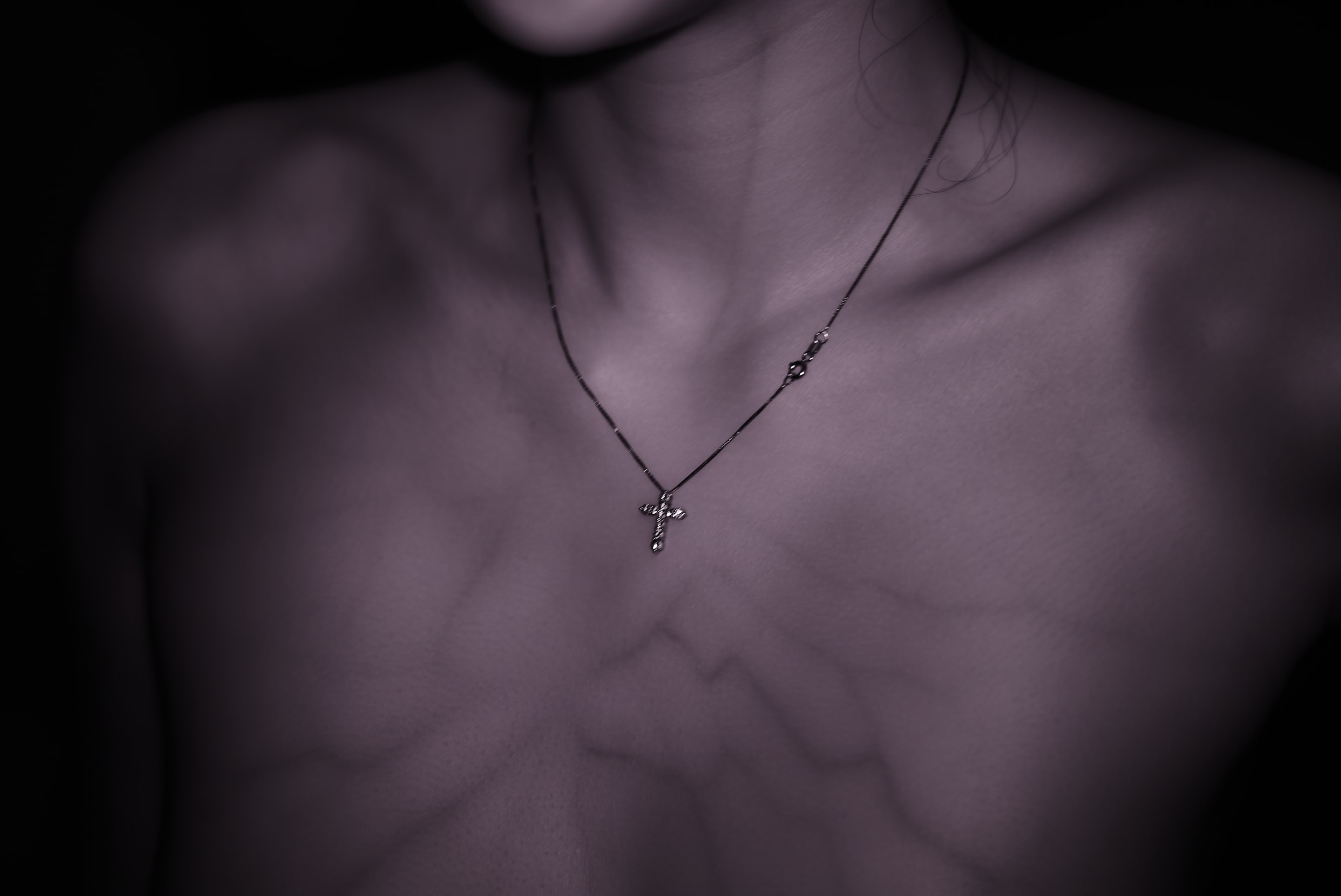 A close-up shot captures the delicate collarbones and neck of a person wearing a thin necklace with a cross pendant. The lighting creates dramatic shadows, highlighting the texture of the skin.