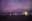 A nighttime beach scene under a vibrant purple sky. Silhouettes of people on the beach contrast with the lights reflecting on the water. blurred out low res