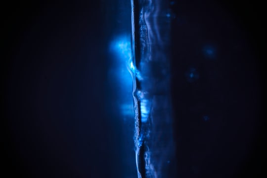 A close-up, abstract shot captures the ethereal luminescence of a dark blue liquid as it cascades down a textured surface. The play of light and shadow creates a sense of depth and mystery.