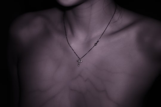 A close-up shot captures the delicate collarbones and neck of a person wearing a thin necklace with a cross pendant. The lighting creates dramatic shadows, highlighting the texture of the skin.