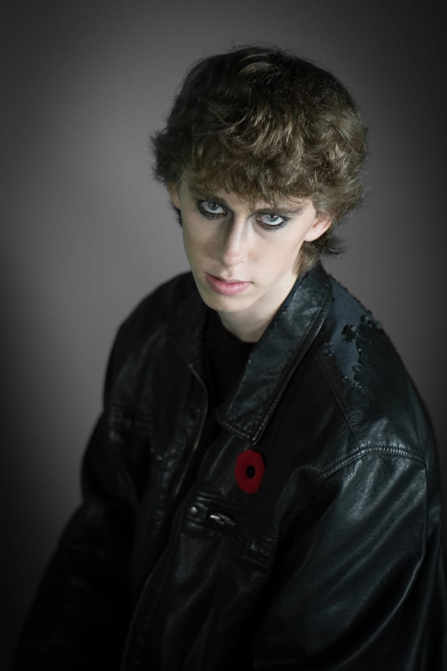 This is a portrait of a person, wearing a black leather jacket with a poppy on it and dark makeup, set against a gray background. The subject is looking directly at the viewer.