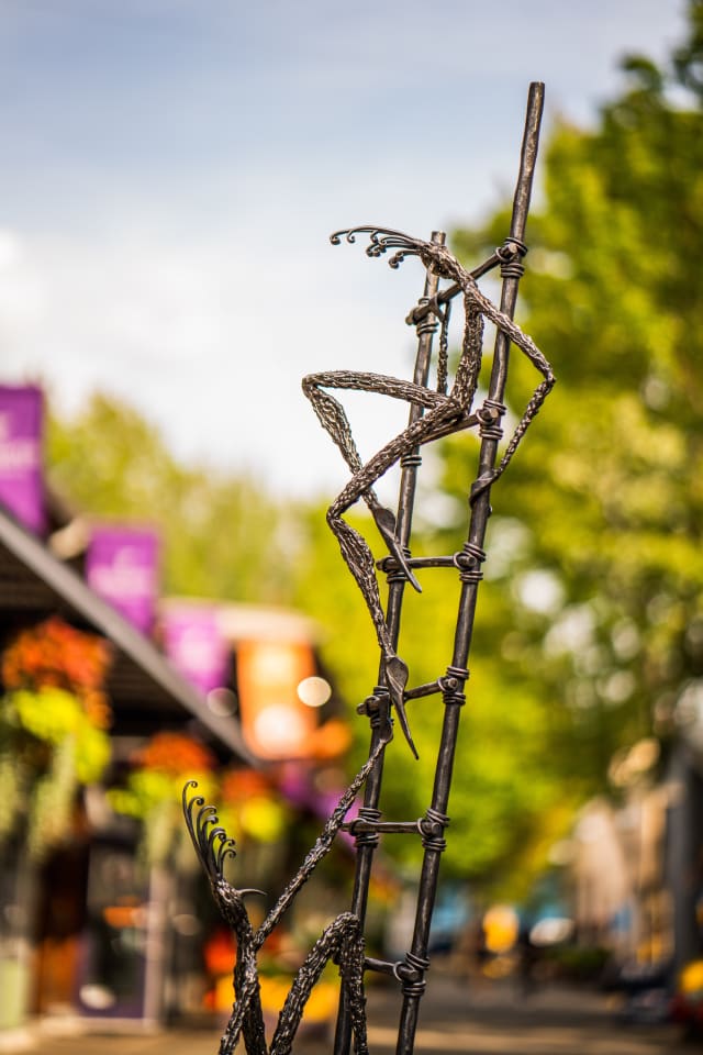 This image showcases an abstract metal sculpture of a figure climbing a ladder. The sculpture is set against a blurred background of greenery and buildings.