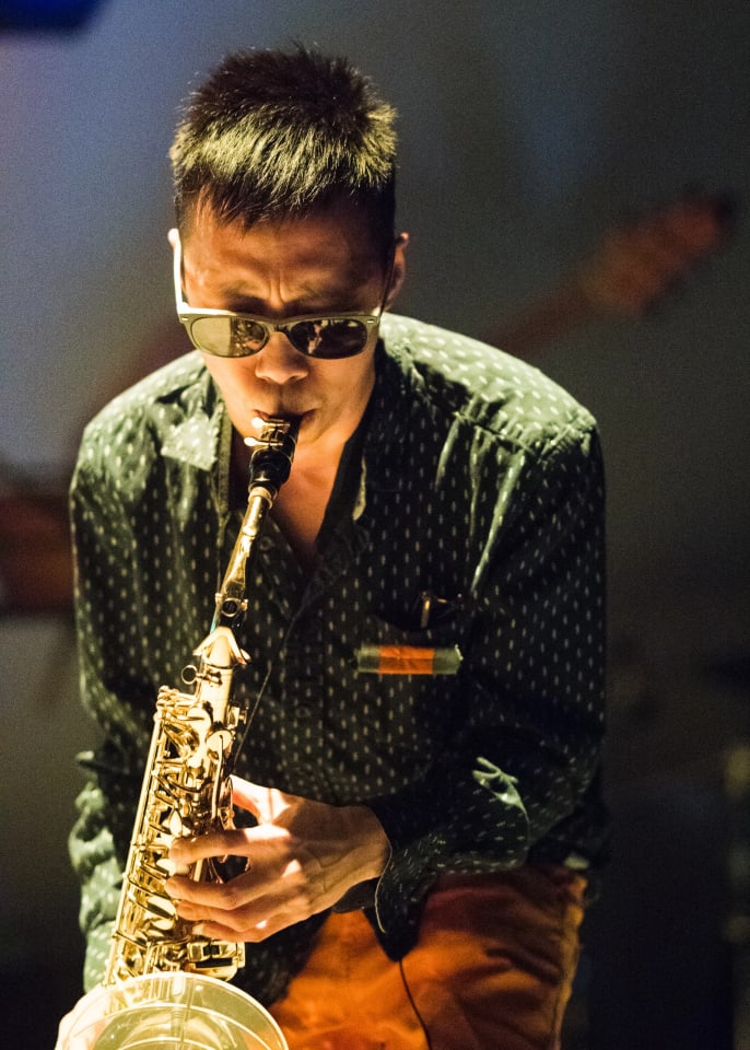 A man in sunglasses plays a saxophone, with his face partially visible. The scene appears to be lit by stage lighting.