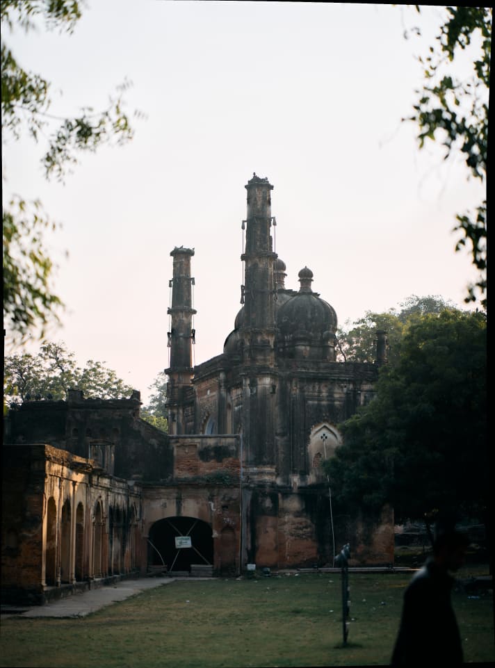 An old building with multiple towers is framed by trees, with a person in the bottom right. The building has a dark color, and some structures like arches can be seen on the sides.