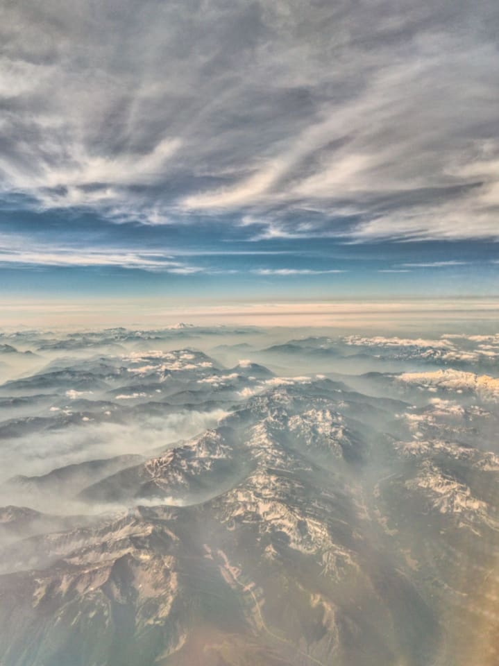 An aerial view of a mountain range covered in snow and mist, under a dramatic cloudy sky. The scene captures the vastness and serenity of nature.
