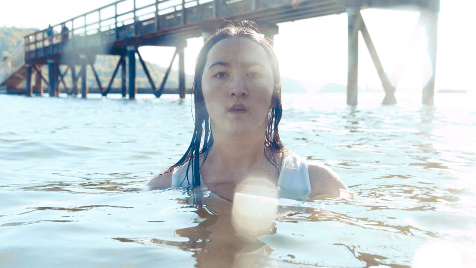 A woman emerges from the water, facing the viewer with a contemplative expression. The scene is bathed in sunlight, with a pier structure visible in the background.