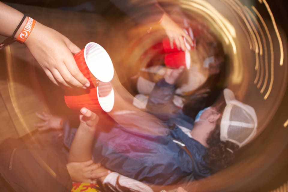 A blurry image featuring hands holding red cups and a person wearing a baseball cap. The overall composition suggests a party or social gathering.