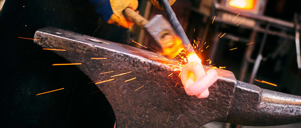 A blacksmith forges metal on an anvil, sparks flying from the impact of a hammer. The image captures the intensity and artistry of traditional metalworking.