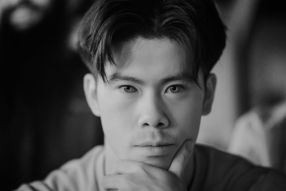 A black and white close-up portrait of a young Asian man. The image focuses on his face, capturing his contemplative gaze.