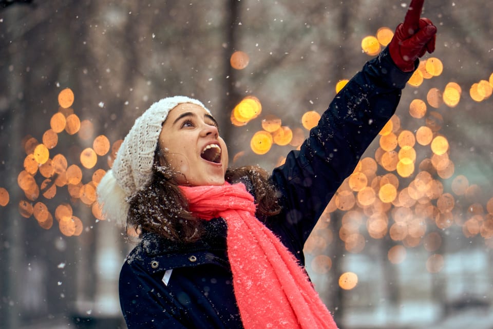 A woman joyfully points upwards while it snows. The background features bokeh lights, enhancing the festive atmosphere.