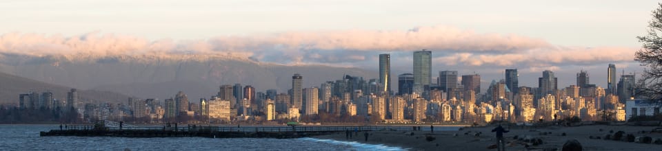 This is a panoramic photograph of a city skyline at sunset, with mountains and clouds in the background, and a beach in the foreground.