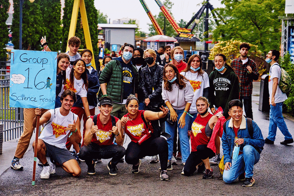 A group of young adults poses for a photo at an amusement park. Most of them are wearing face masks, and holding a sign that says "Group 16 Science Frosh."