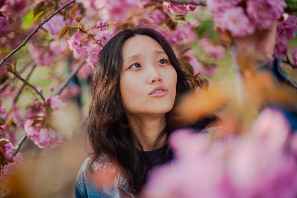 A young woman gazes upward amidst a vibrant display of pink cherry blossoms, bathed in soft, natural light. The composition highlights the interplay of the subject's expression and the surrounding floral beauty.