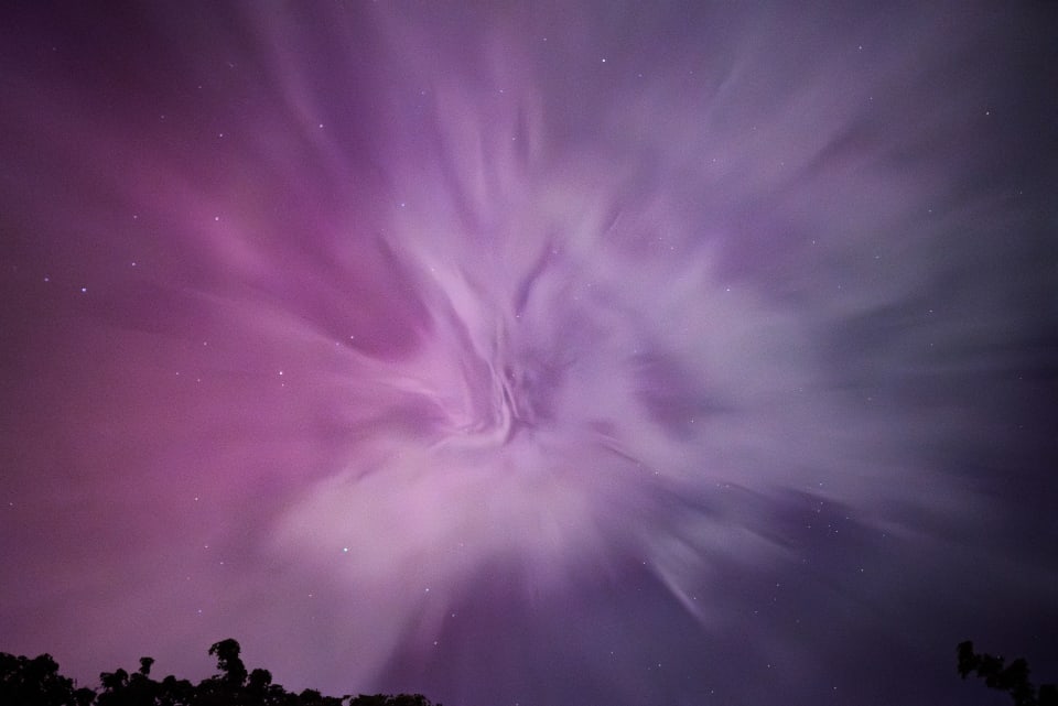 A vivid, ethereal capture of a night sky. The image showcases swirling clouds and distant stars set against a mesmerizing palette of purple and pink.