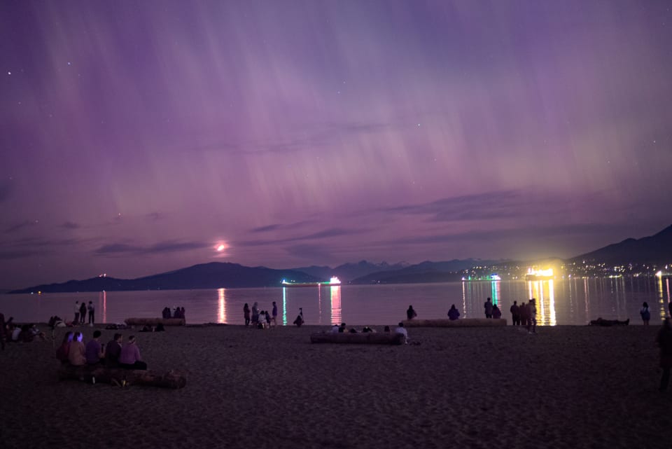 A nighttime beach scene under a vibrant purple sky. Silhouettes of people on the beach contrast with the lights reflecting on the water.