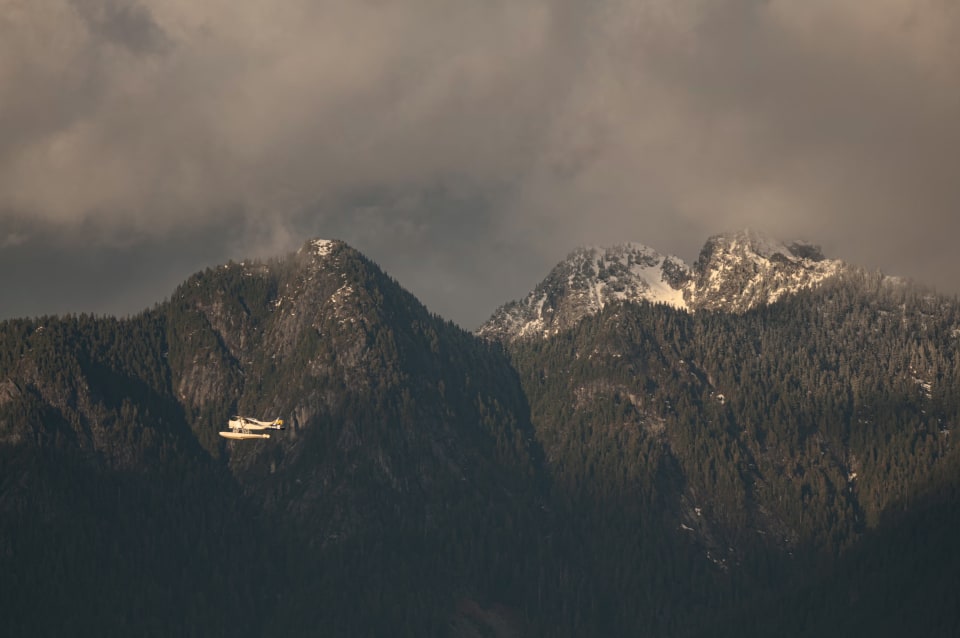 A seaplane soars against a backdrop of towering, snow-dusted mountains. The scene is enveloped in the soft light of a cloudy day.