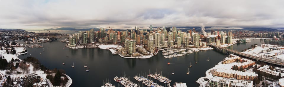 An aerial panoramic view of a city skyline on a cloudy day. The city has tall buildings and is situated near a body of water with boats.
