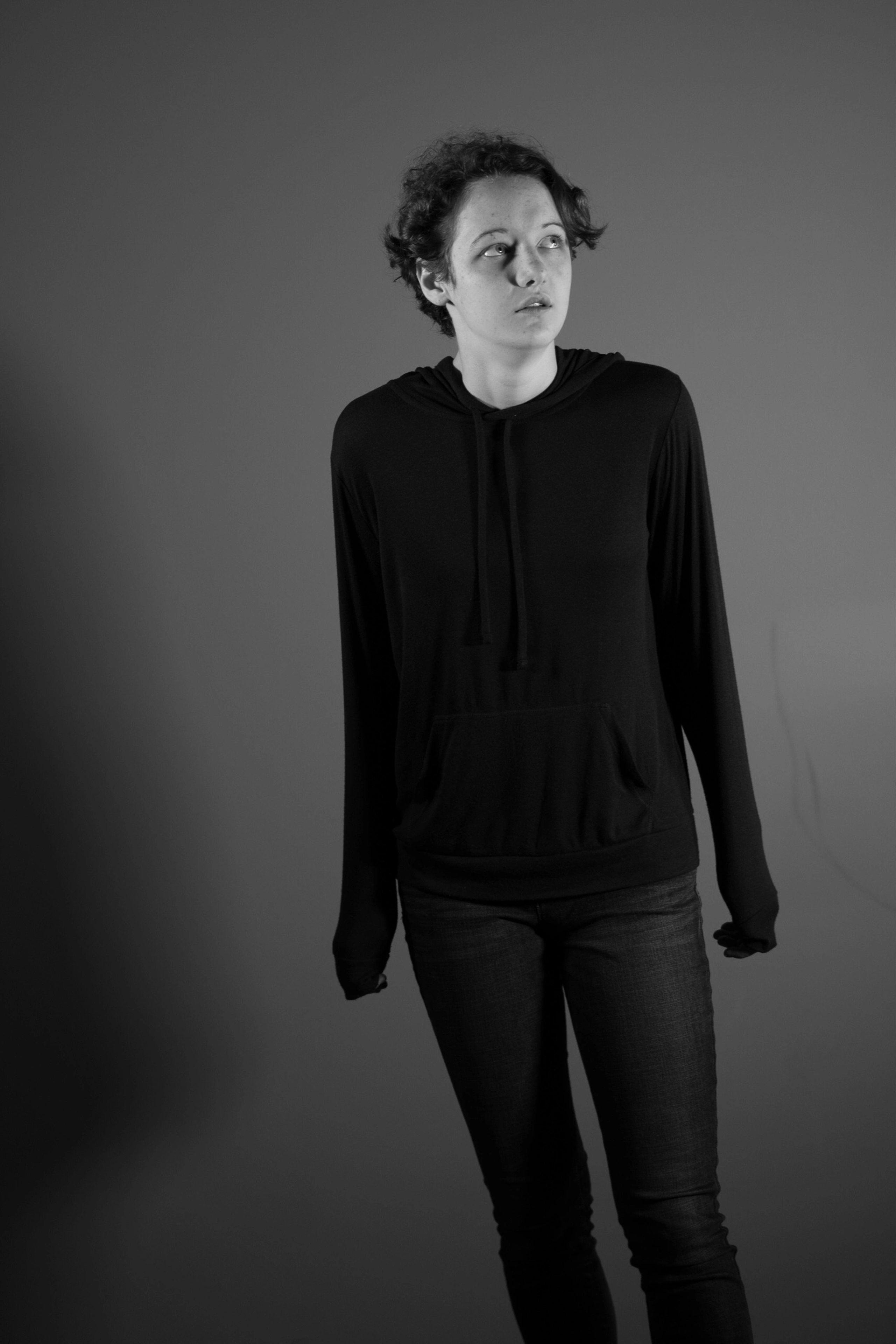 A black-and-white studio portrait of a person in a hoodie and jeans looking upward.