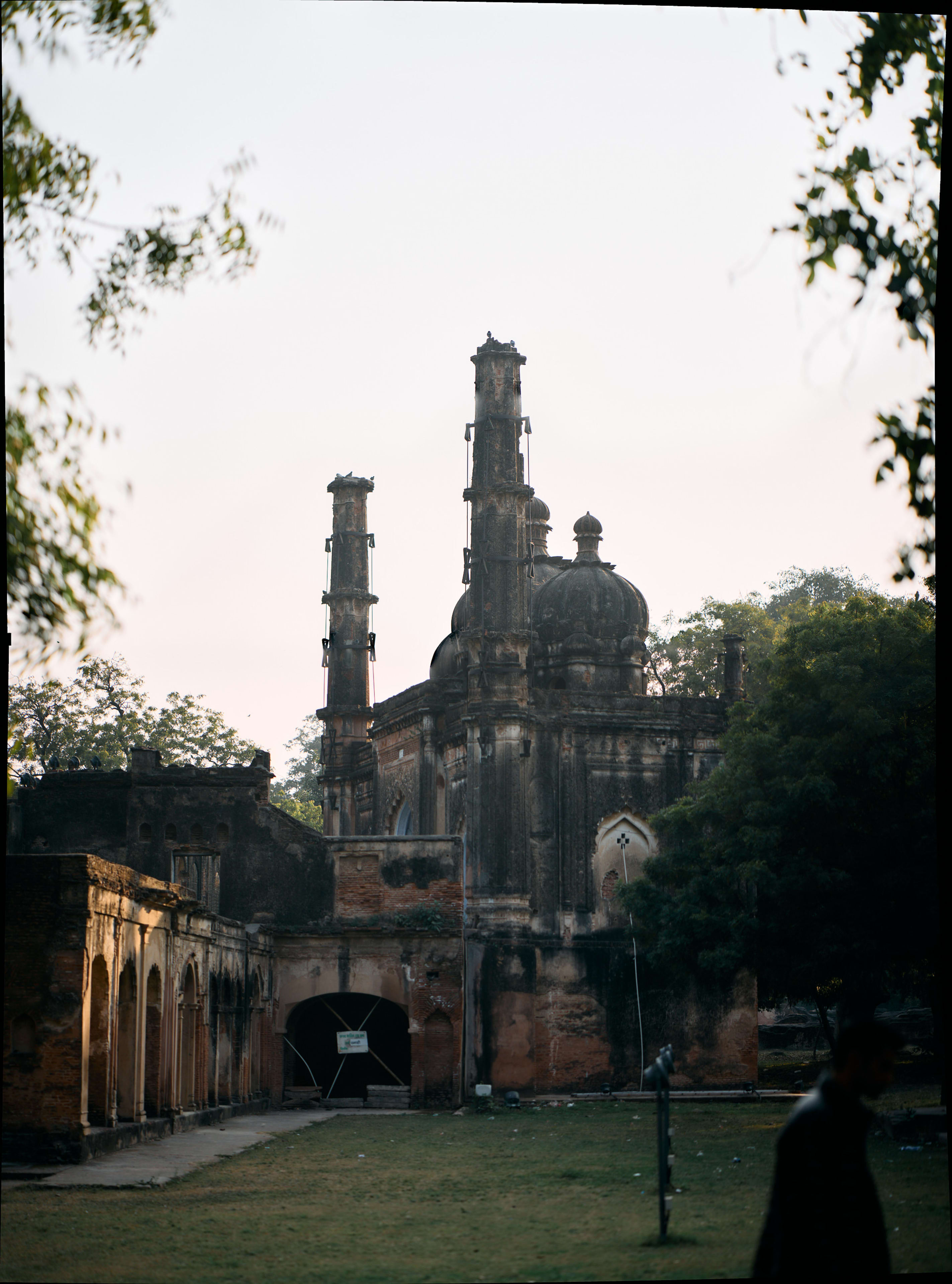 An old building with multiple towers is framed by trees, with a person in the bottom right. The building has a dark color, and some structures like arches can be seen on the sides.