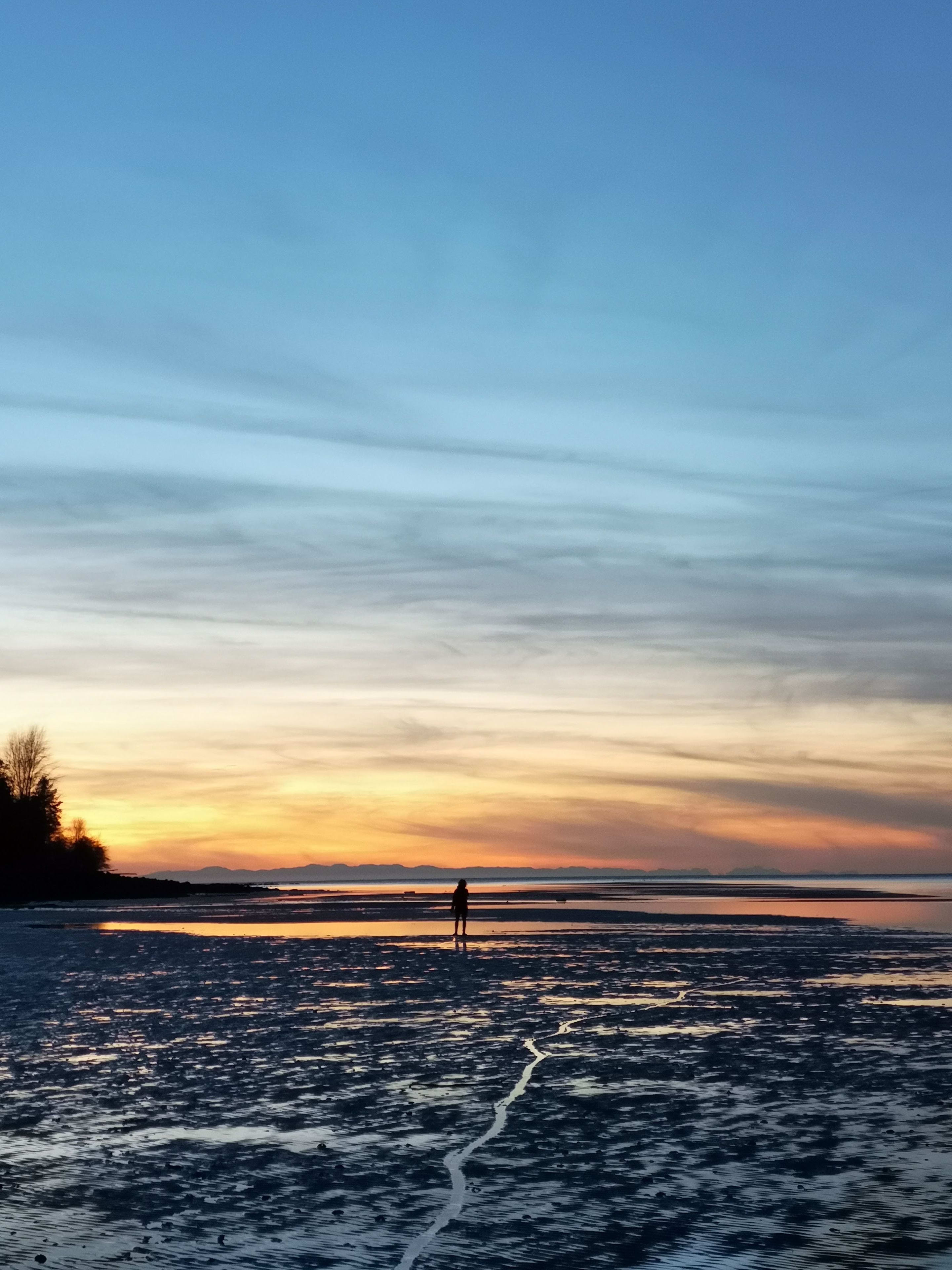 A silhouetted figure stands on wet sand at sunset while the sky reflects in the water.