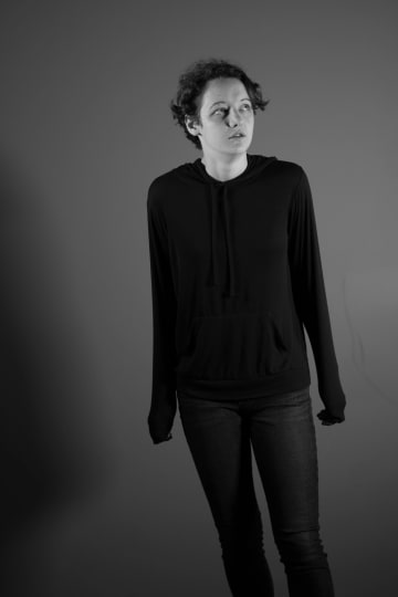 A black-and-white studio portrait of a person in a hoodie and jeans looking upward.