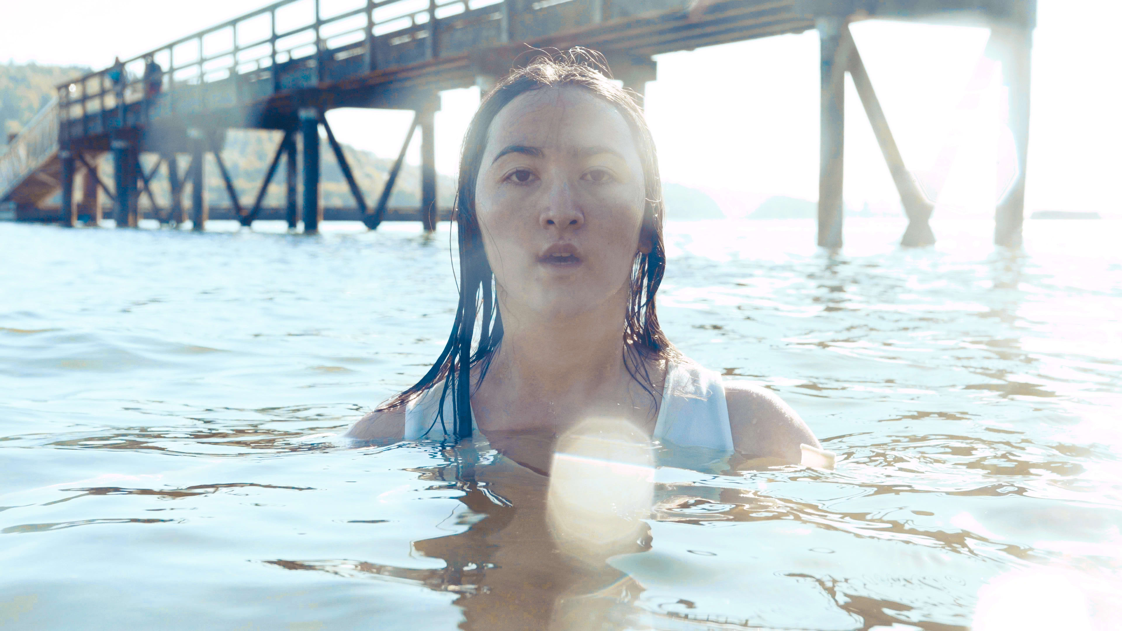 A woman rises from the water in bright sunlight, with a pier structure behind her.