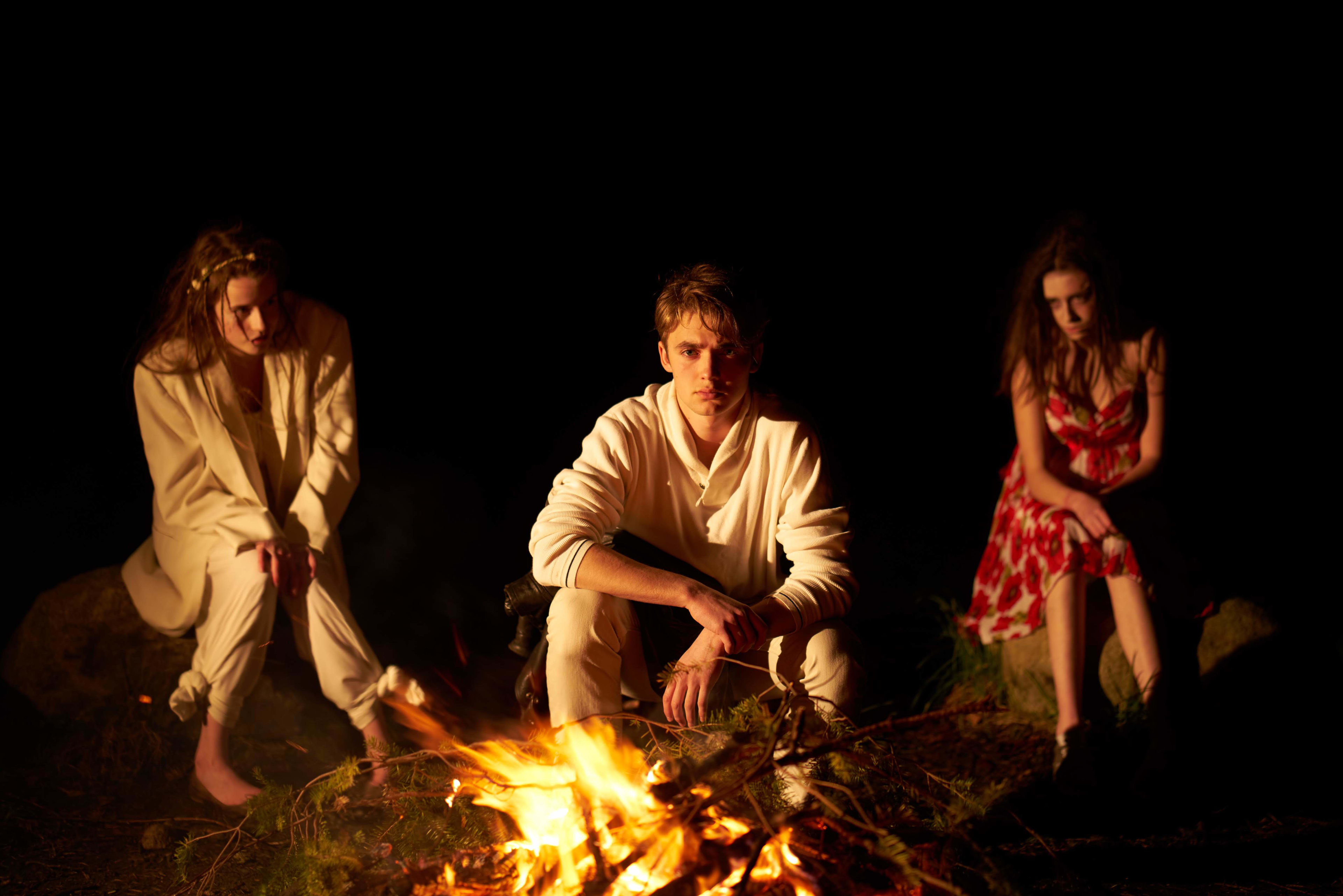 Three people sit around a campfire at night. The fire does most of the lighting.