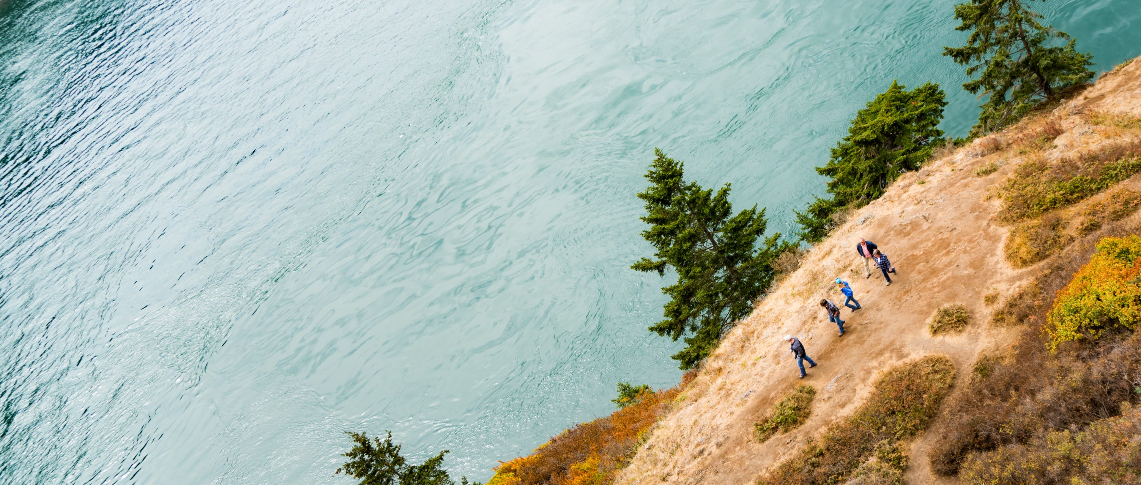 A group walks along a hillside above open water.