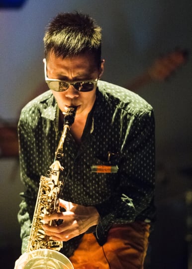 A man in sunglasses plays saxophone under stage light.