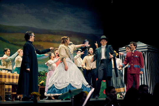 A cast in period costume stands on stage under green light.