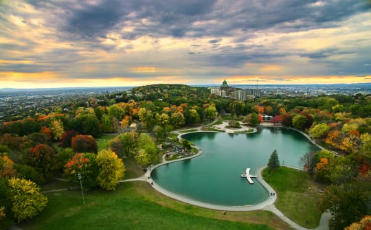An aerial view of a park during autumn, featuring a lake surrounded by trees with colorful foliage and a sunset in the background.