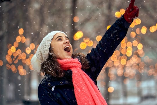 A woman points upward while snow falls and out-of-focus lights fill the background.