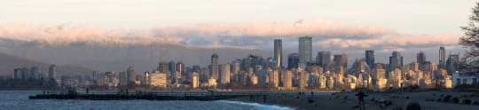 This is a panoramic photograph of a city skyline at sunset, with mountains and clouds in the background, and a beach in the foreground.