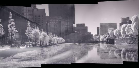 A black and white photograph of a cityscape featuring buildings, trees, and a body of water. The image has a high contrast and a sense of stillness.
