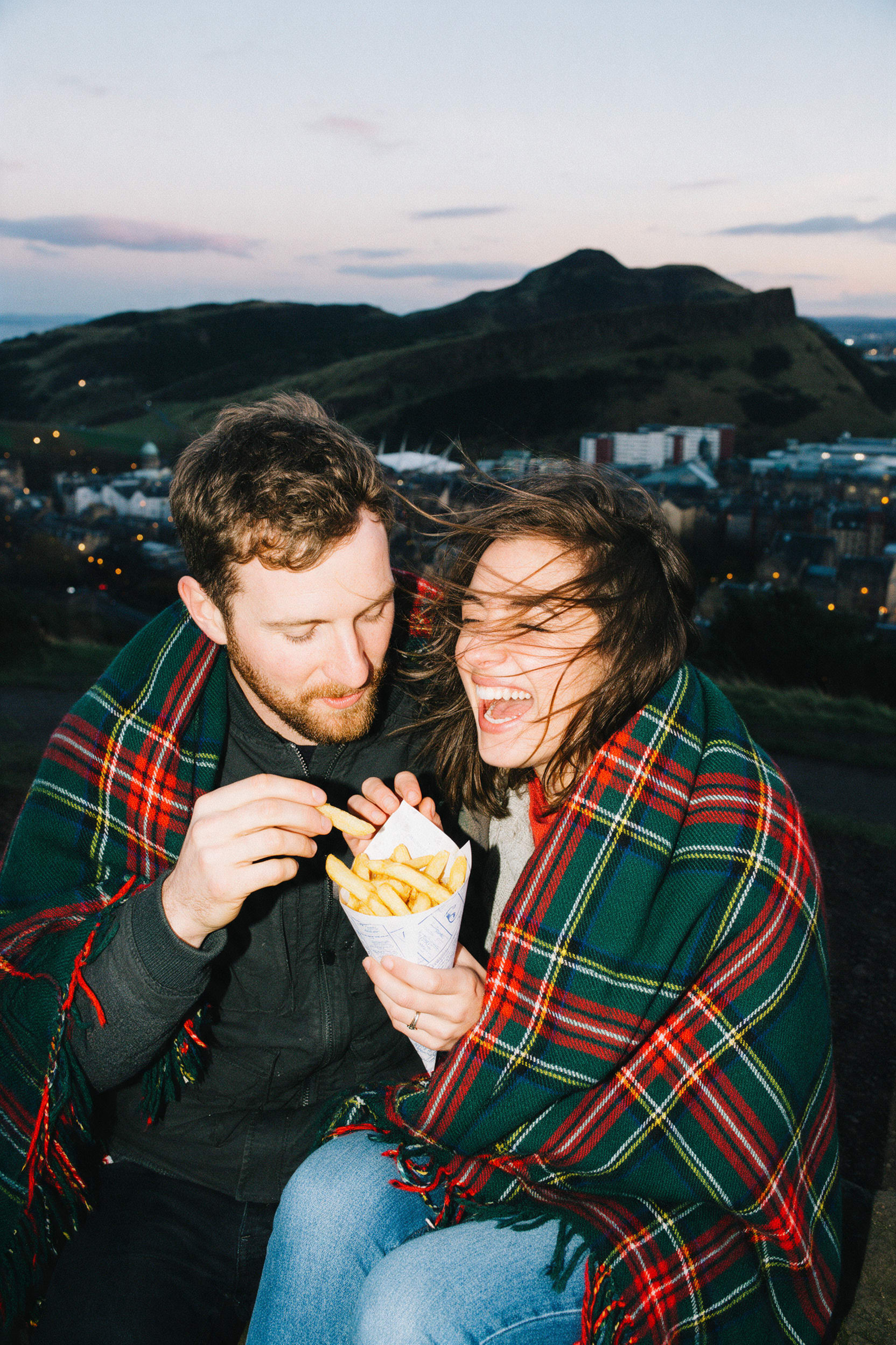 A couple wrapped in a tartan blanket sharing a cone of chips above a city at dusk
