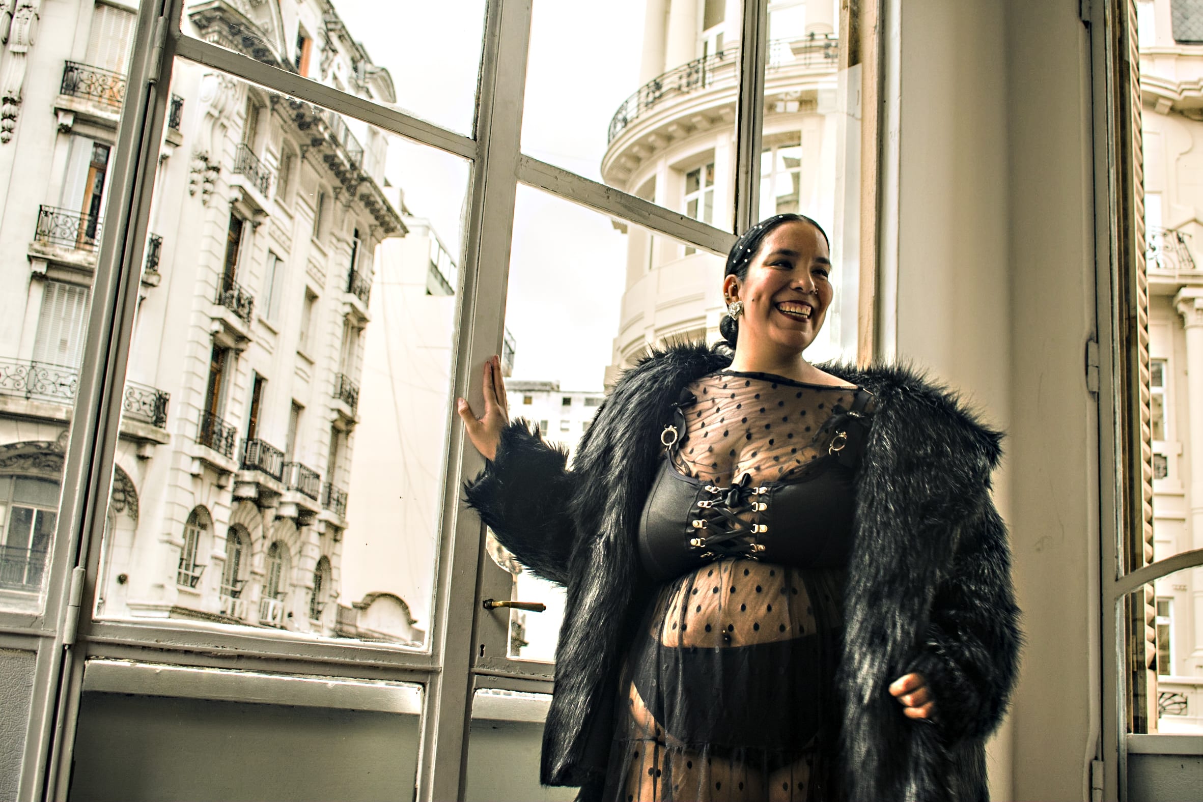 Mujer sonriente vistiendo un abrigo de piel sintética negra y lencería con transparencias, mirando por un gran ventanal hacia edificios clásicos. Sesión boudoir urbana con luz natural.
