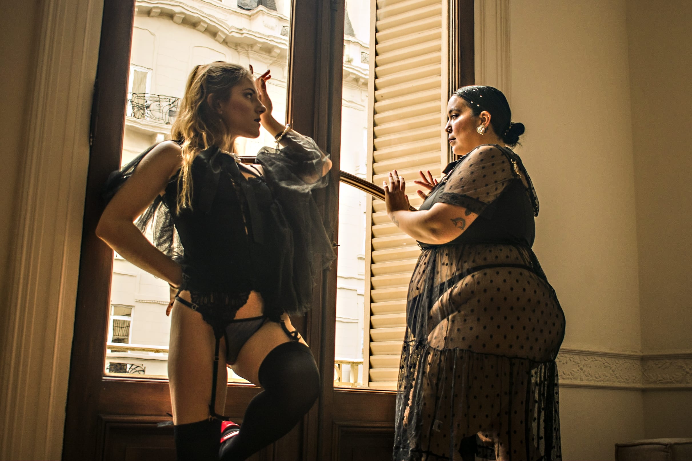Dos mujeres posando juntas frente a un gran ventanal antiguo iluminado por luz cálida, luciendo lencería negra y prendas translúcidas. Fotografía boudoir conceptual en pareja.