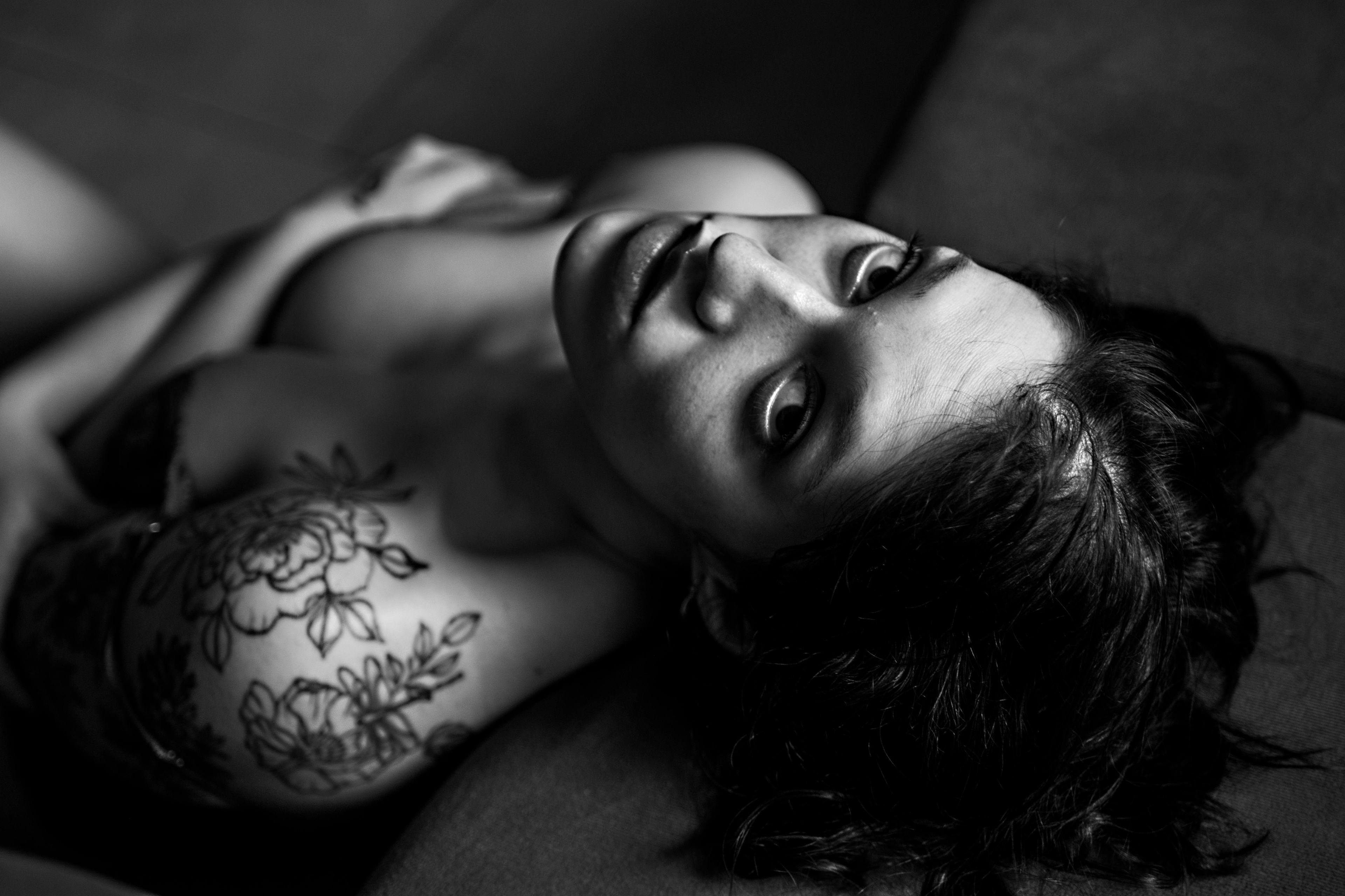 Retrato íntimo en blanco y negro de una mujer recostada mirando a la cámara, destacando un tatuaje floral en el hombro. Fotografía boudoir de alta gama en La Plata por Vanguardia.