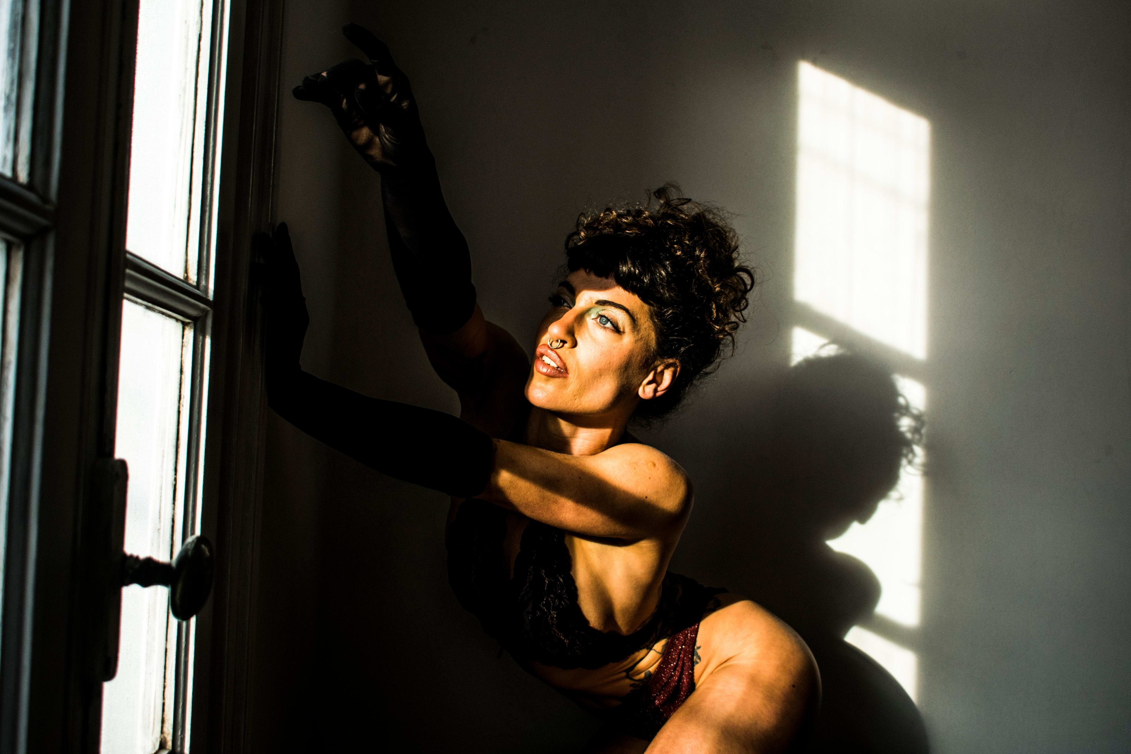 Mujer posando de forma teatral junto a una ventana, iluminada por un rayo de sol que crea sombras dramáticas. Viste guantes largos negros en una sesión de arte boudoir conceptual.