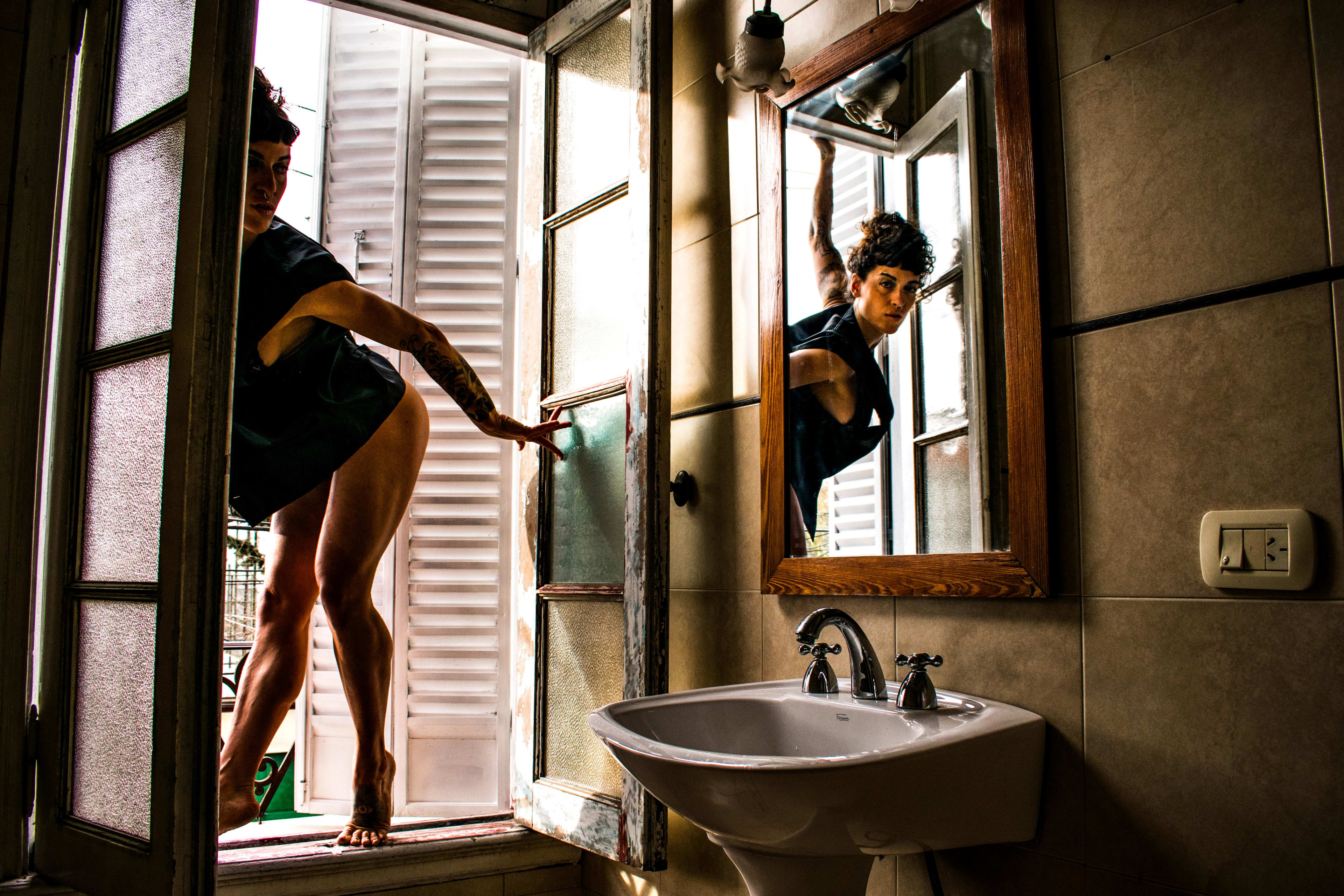 Composición fotográfica creativa mostrando el reflejo en un espejo de baño de una mujer posando dinámicamente en el marco de una puerta. Sesión boudoir inmersiva y conceptual.