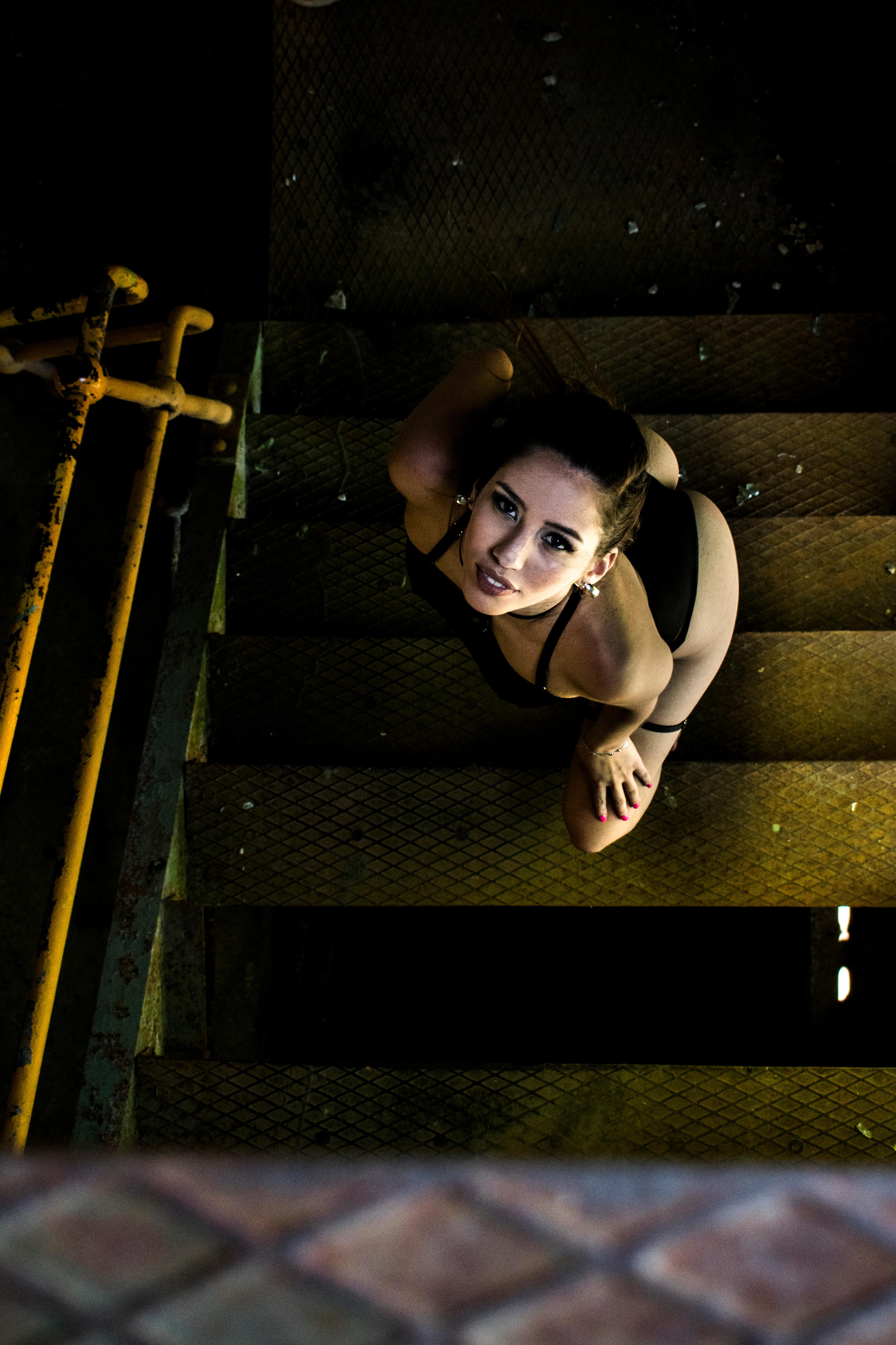 Retrato en picado de una mujer posando en lencería negra sobre una escalera metálica industrial. Sesión fotográfica boudoir urbana y performativa por Vanguardia.