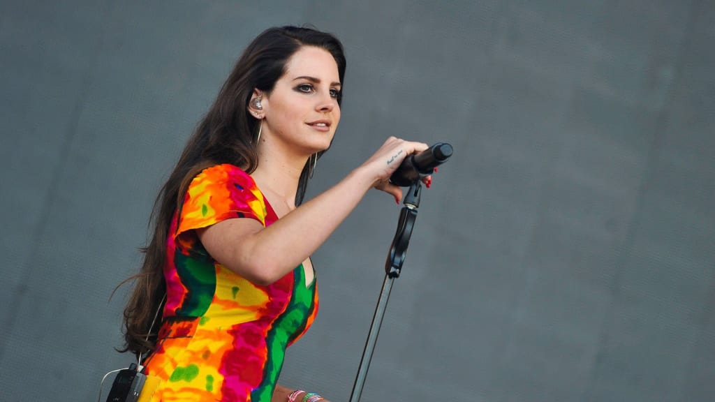 Singer performing on stage wearing vibrant multicolored outfit, holding microphone against gray background with focused expre