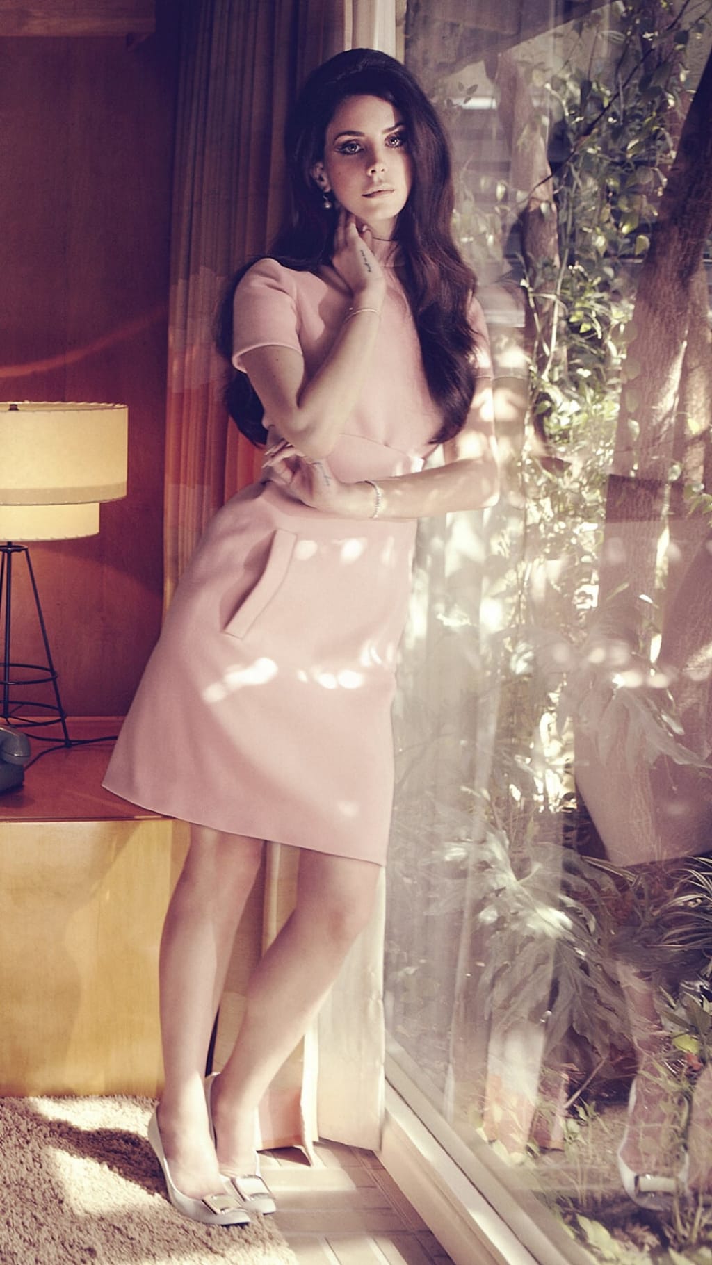Woman in pink dress standing by illuminated plants and warm lamp lighting, vintage aesthetic with moody atmospheric ambiance