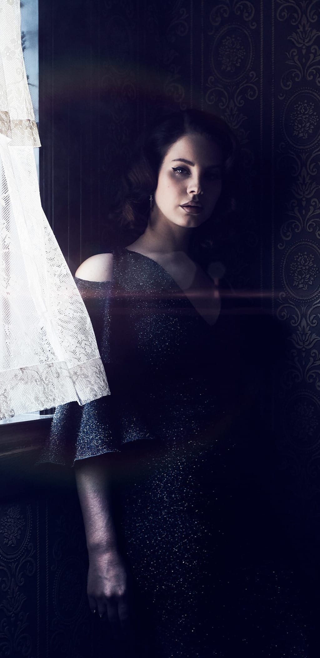 Woman in sparkly dark dress posed against ornate patterned backdrop with white sheer fabric, atmospheric moody lighting and i