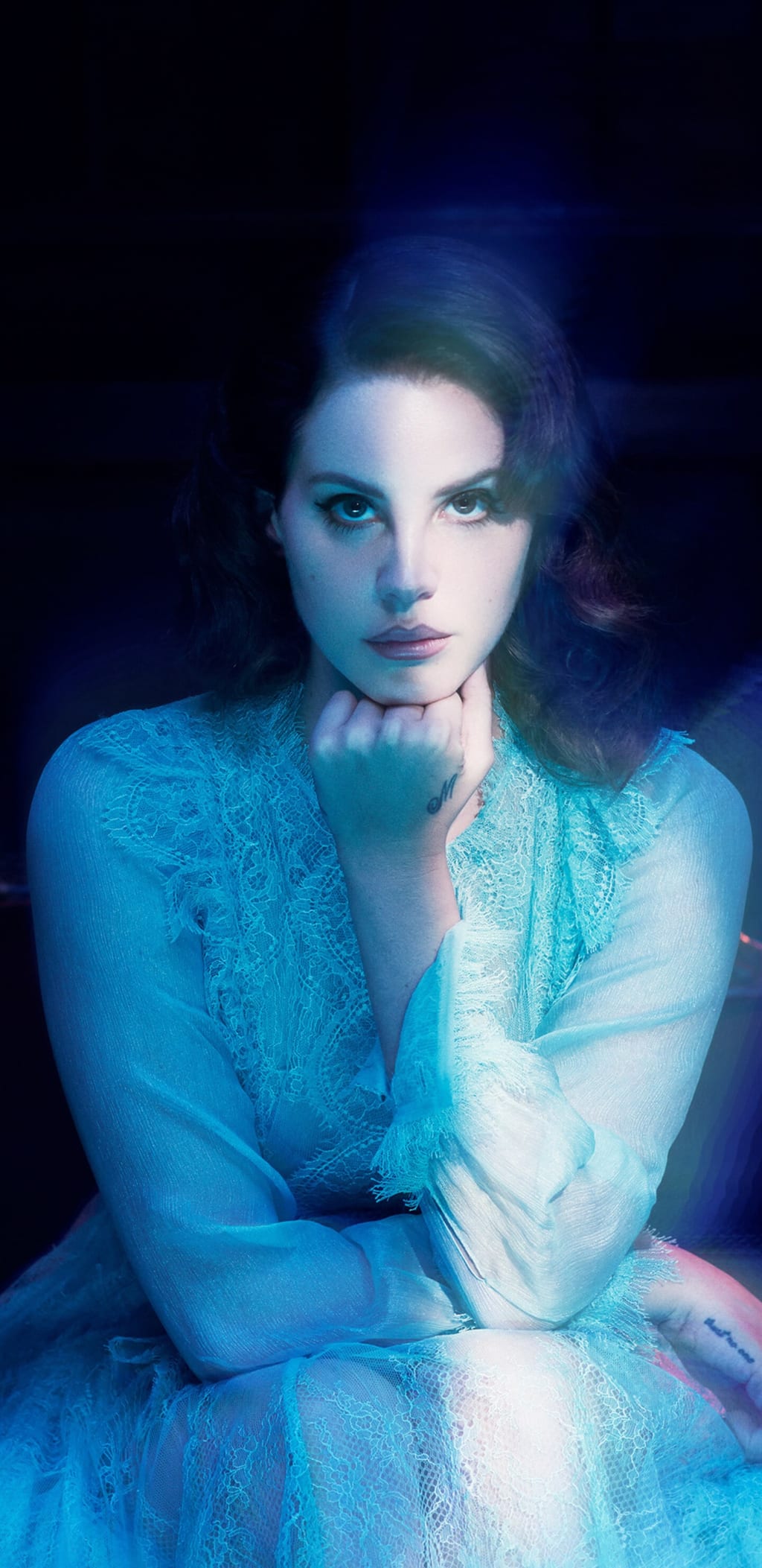 Woman with dark hair in ethereal blue-tinted lighting, wearing delicate lace clothing, gazing pensively with hand near chin a